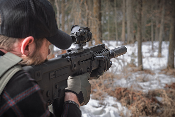 Over-the-shoulder view of a shooter aiming a compact rifle equipped with an Off Grid Suppressors Trident TI titanium suppressor and red dot optic in a snowy wooded environment.