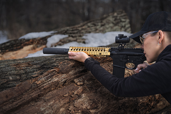 Person wearing safety glasses and a black cap aims an AR-style rifle with a gold handguard and an Off Grid Suppressors suppressor while resting the rifle on a fallen log in a snowy wooded area. The image highlights the suppressor's lightweight design and use in an outdoor shooting environment.