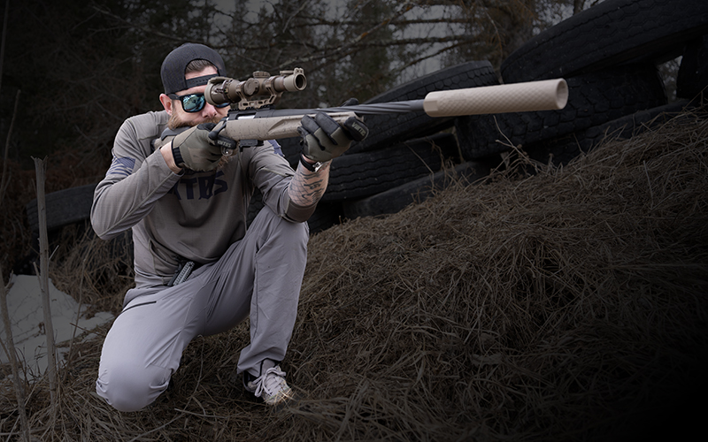 OGS-About-Us-2 Person wearing gloves, sunglasses, and a gray outfit kneels while aiming a bolt-action rifle equipped with an Off Grid Suppressors Trident TI titanium suppressor and scope. Stacked tires and a dirt berm form the backdrop at an outdoor shooting range.