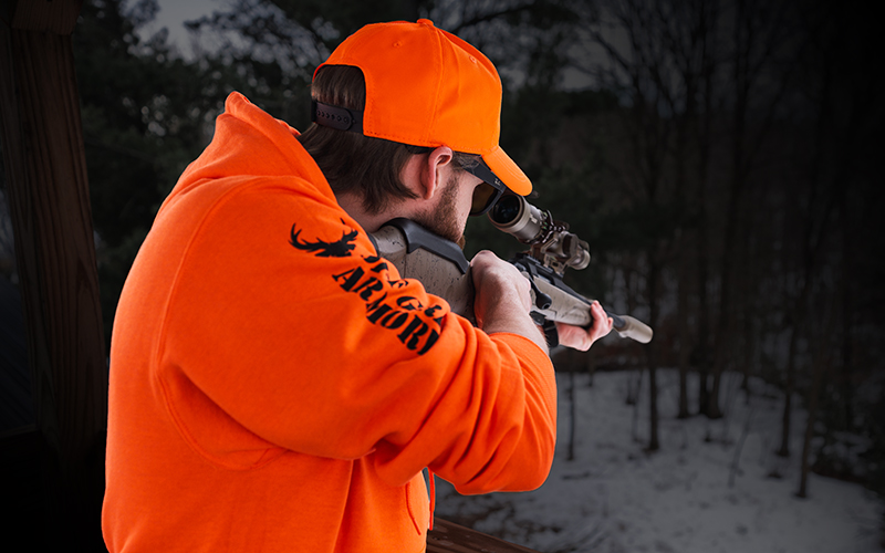 OGS-About-Us-3-Trident Rear view of a person in a bright orange hoodie and cap aiming a scoped bolt-action rifle equipped with an Off Grid Suppressors Trident TI titanium suppressor from a covered outdoor shooting platform overlooking a wooded, snow-covered landscape.