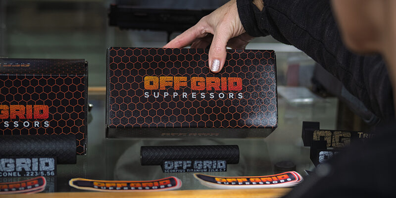 OGS-About-Us-1 Customer holding an Off Grid Suppressors product box over a display case featuring branded suppressors, product decals, and retail packaging. The display highlights Off Grid Suppressors branding and products available for purchase through authorized dealers.